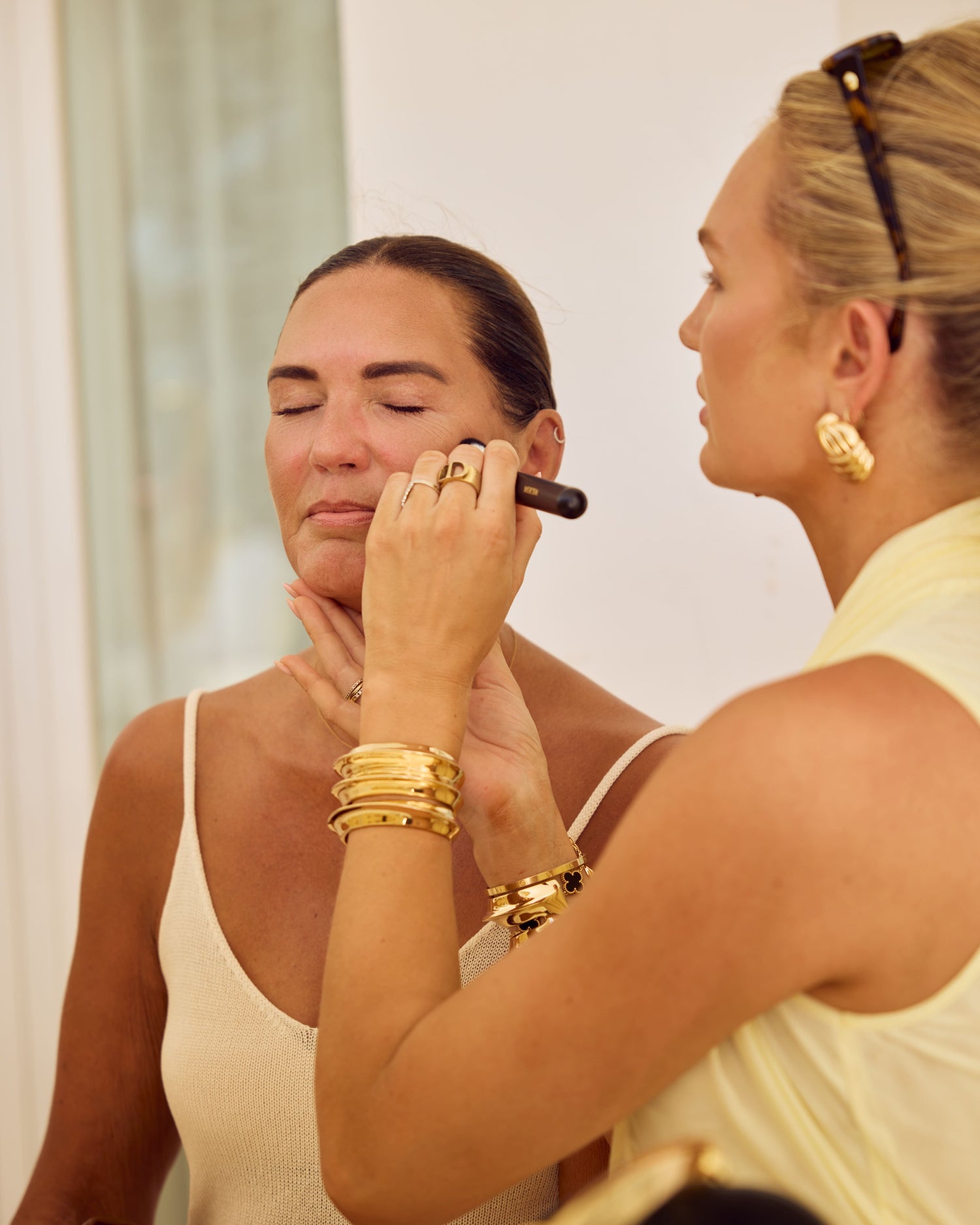 Woman applying makeup to another woman's face in a mirror.