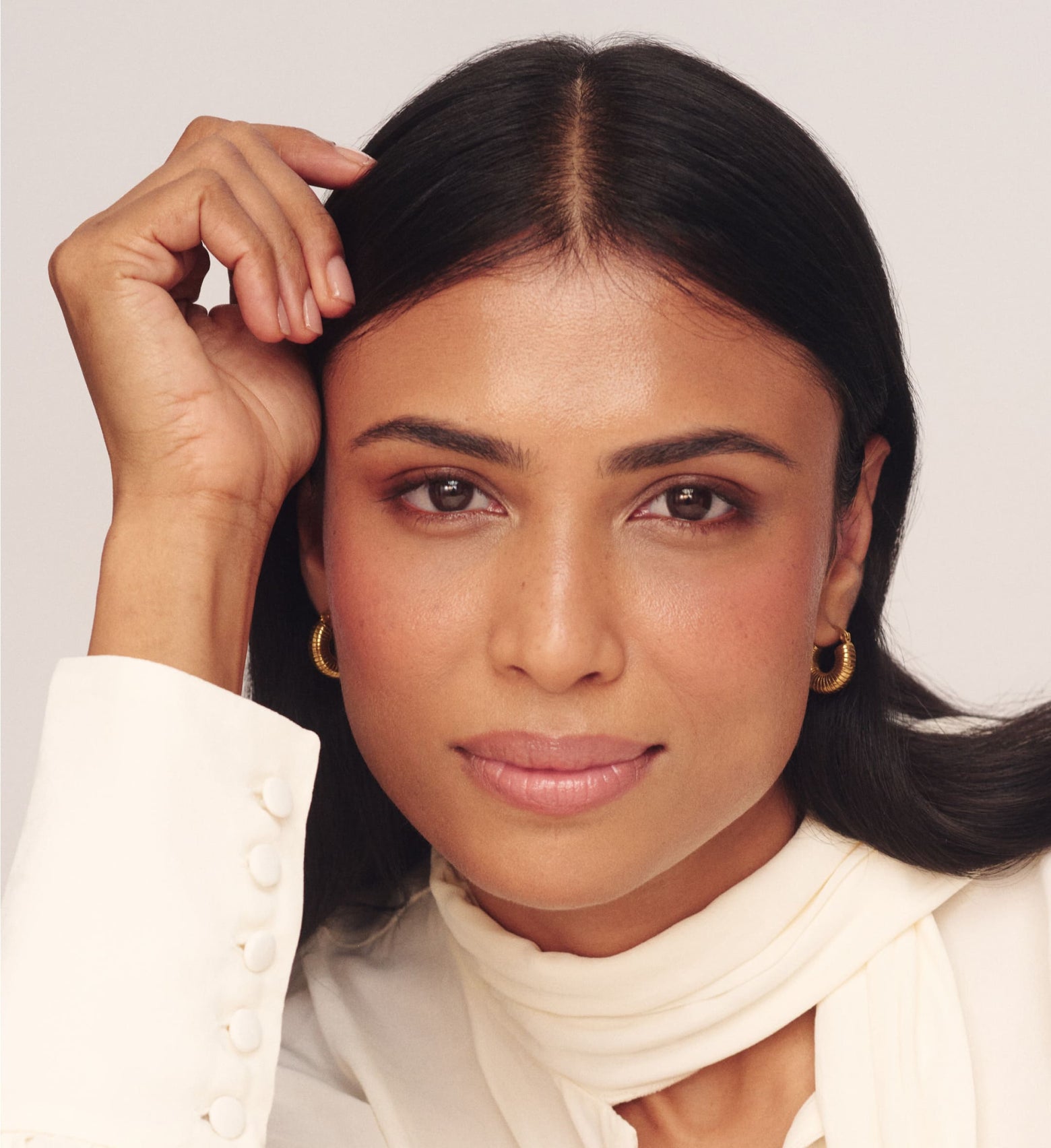 Woman with dark hair and earrings wearing a white top against a light background
